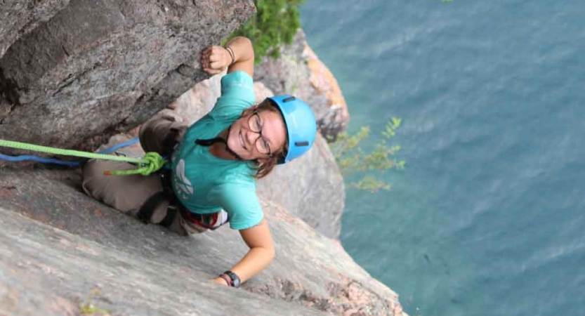 a person who is rock climbing above a body of water smiles at the camera